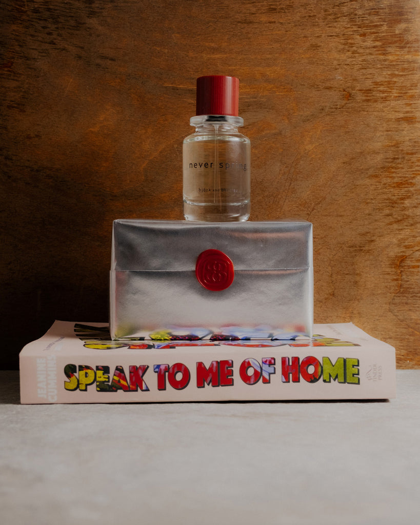 Never spring Perfume bottle, silver wrapped box with red seal, and book titled 'Speak to Me of Home' on a wooden surface.