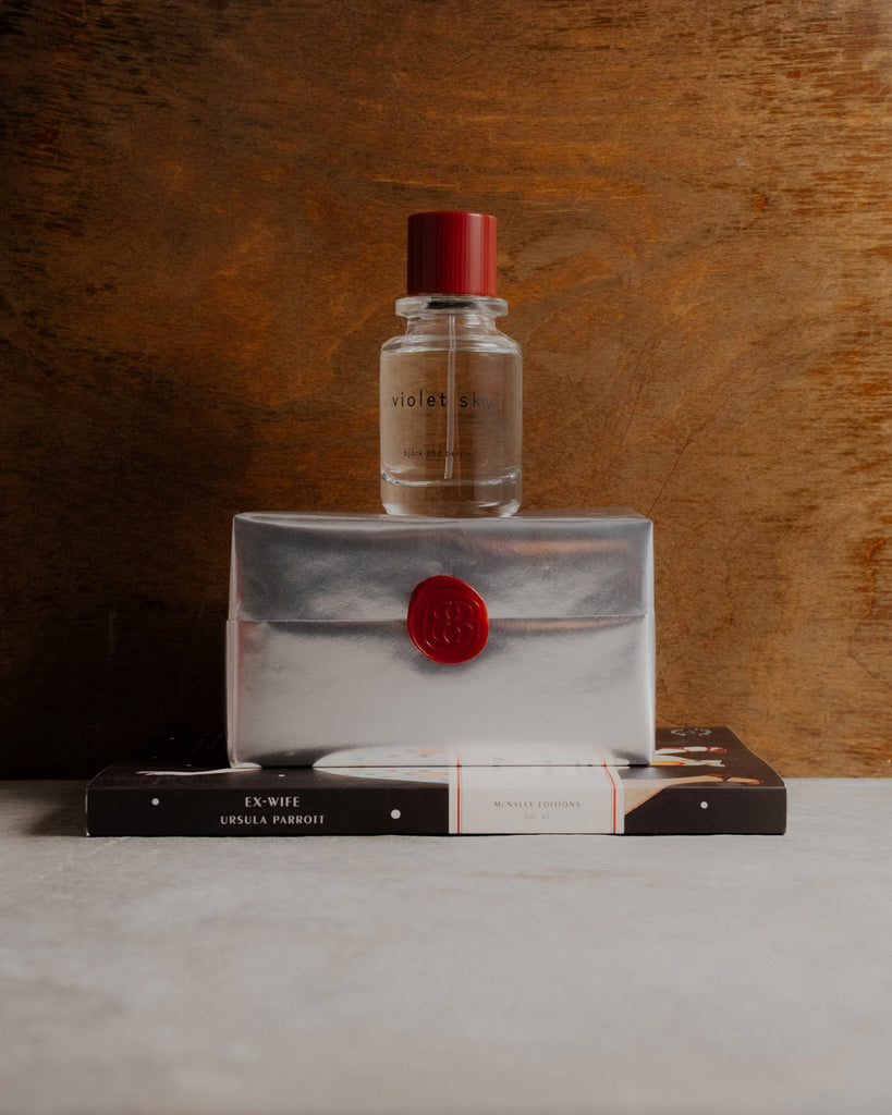 Violet sky edp bottle on a silver wrapped perfume box with a red seal, placed on top of a book titled 'Ex-wife'.