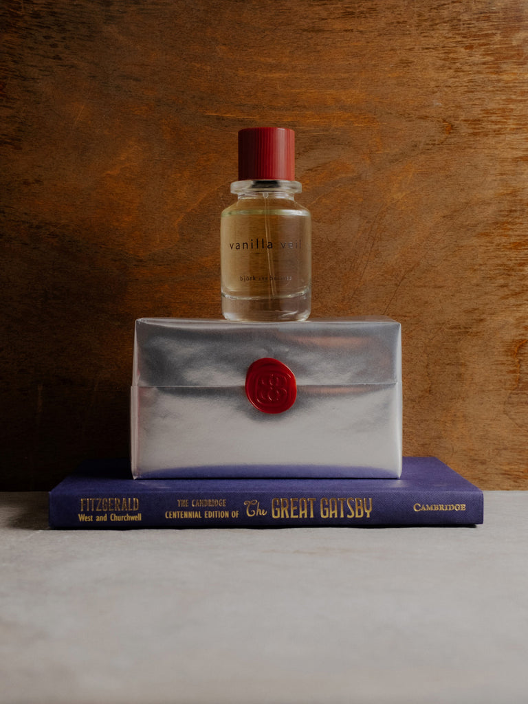 Vanilla veil edp bottle on a silver wrapped perfume box with a red seal, placed on top of a book titled 'The Great Gatsby'.