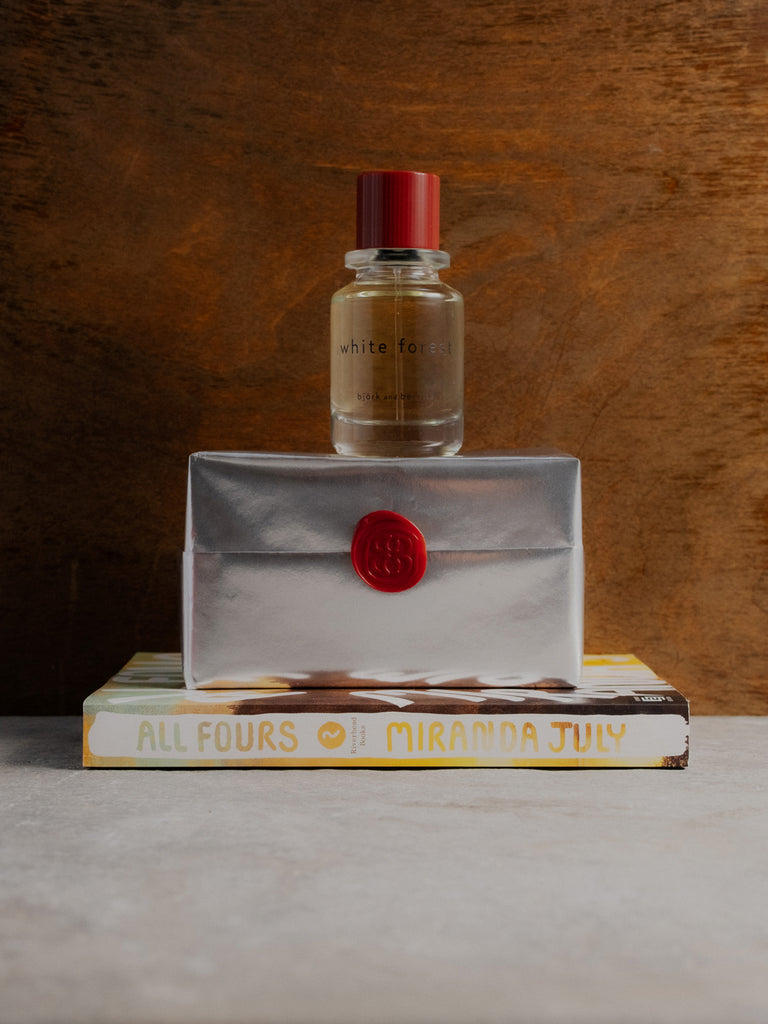 White forest edp bottle on a silver wrapped perfume box with a red seal, placed on top of a book titled 'All fours'.